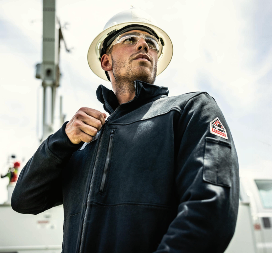 Person wearing a hard hat and black jacket with a logo, standing outdoors.